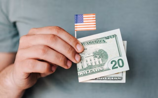 Dłoń trzyma małą flagę USA oraz banknoty dwudziestodolarowe na szarym tle