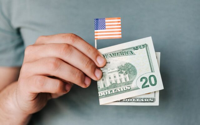 Dłoń trzyma małą flagę USA oraz banknoty dwudziestodolarowe na szarym tle