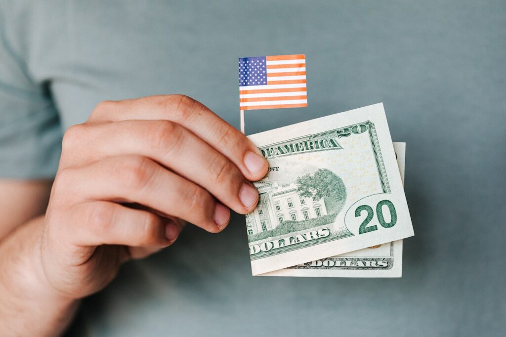 Dłoń trzyma małą flagę USA oraz banknoty dwudziestodolarowe na szarym tle