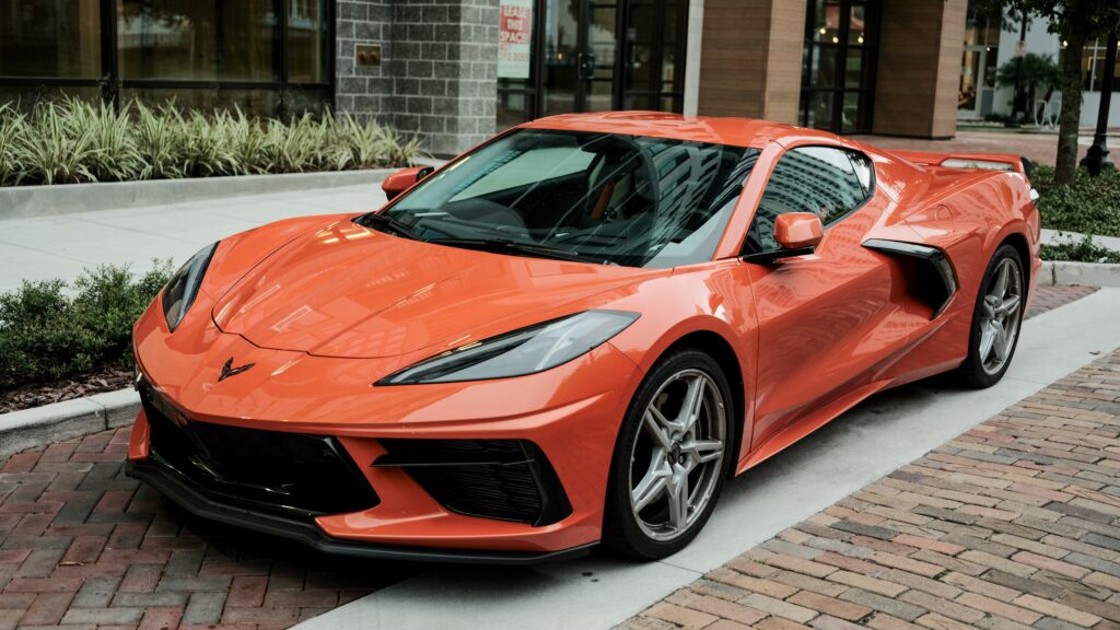 Pomarańczowe Chevrolet Corvette C8 zaparkowane na chodniku przy nowoczesnym budynku, widok z przodu i boku