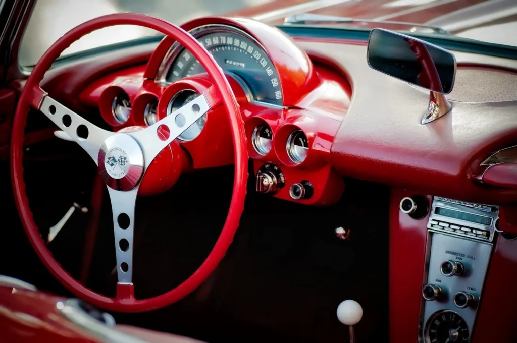 Kokpit klasycznej Corvette z lat 60., z czerwonym wnętrzem, metalową kierownicą z logo Chevrolet Corvette i stylową deską rozdzielczą w stylu retro.