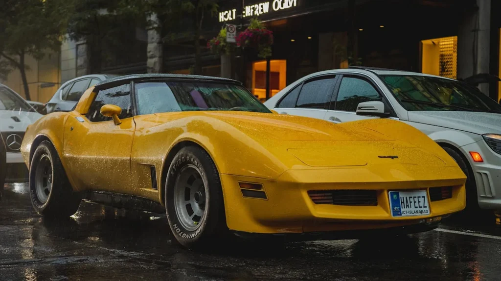 Klasyczna żółta Corvette z USA z lat 70. zaparkowana na mokrej ulicy, z szerokimi oponami i agresywną sylwetką muscle cara.