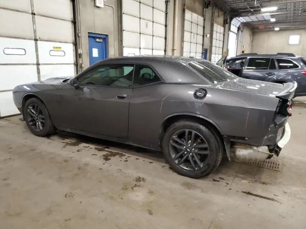 Szary Dodge Challenger coupe z lewej tylnej strony, widoczne uszkodzenie zderzaka w hali warsztatowej.