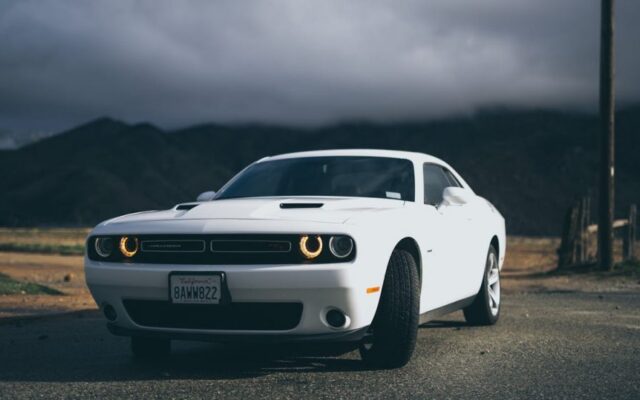 Biały Dodge Challenger z włączonymi światłami stoi na drodze przed ciemnymi górami i burzowym niebem
