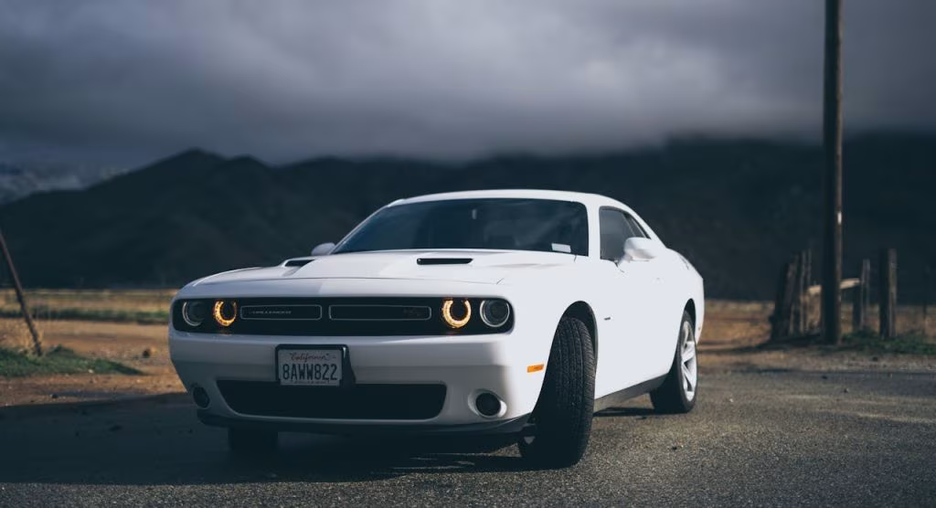 Biały Dodge Challenger z włączonymi światłami stoi na drodze przed ciemnymi górami i burzowym niebem