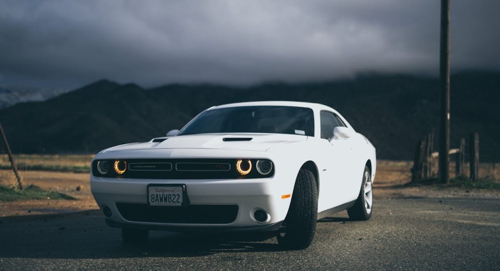 Biały Dodge Challenger z włączonymi światłami stoi na drodze przed ciemnymi górami i burzowym niebem
