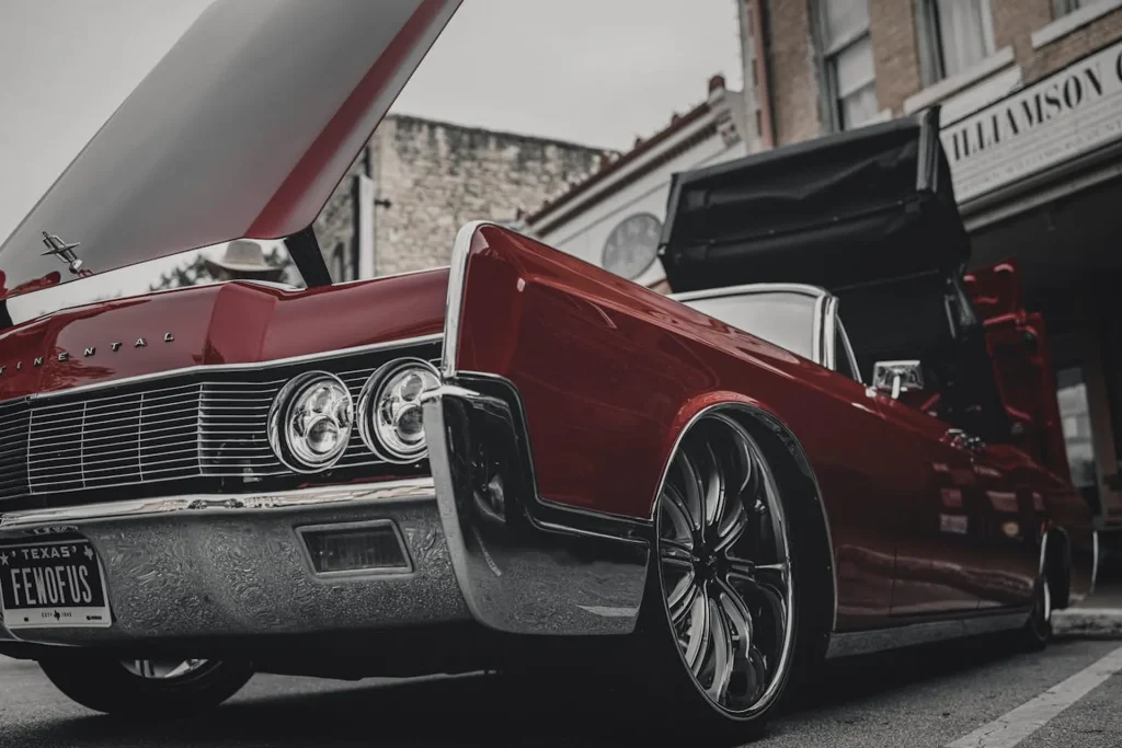 Zdjęcie przedstawia klasyczny, czerwony samochód Lincoln Continental z lat 60-tych, zaparkowany na ulicy. Auto ma charakterystyczne, duże reflektory i chromowane detale. Pojazd ma otwarte drzwi i maskę, ukazując wnętrze i silnik. W tle widać budynki miejskie, co nadaje scenie klimatycznego, retro wyglądu.