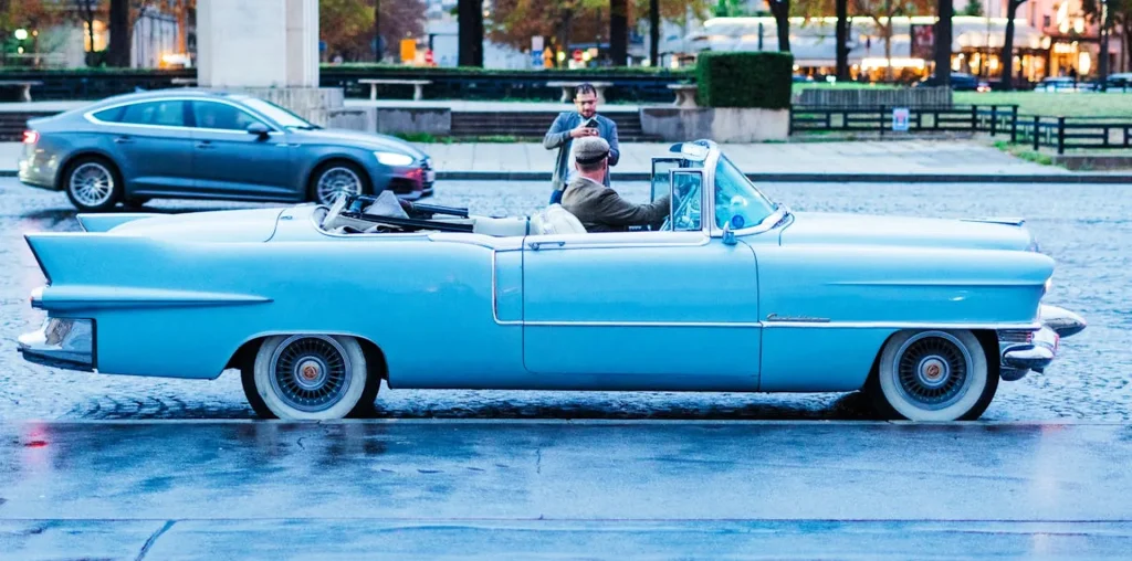Na zdjęciu widzimy klasyczny samochód Cadillac Eldorado w jasnoniebieskim kolorze, będący kabrioletem. Pojazd jest zaparkowany na mokrej ulicy, a za kierownicą siedzi mężczyzna w kaszkiecie, ubrany elegancko, co dodaje scenie retro charakteru. Samochód ma charakterystyczne dla lat 50-tych duże płetwy na tylnych błotnikach i eleganckie, chromowane detale. W tle widać inne pojazdy oraz ludzi, co sugeruje, że zdjęcie zostało zrobione w miejskim otoczeniu, prawdopodobnie w centrum miasta. Refleksy świateł ulicznych na mokrej nawierzchni dodają zdjęciu dynamiki i klimatycznego nastroju.