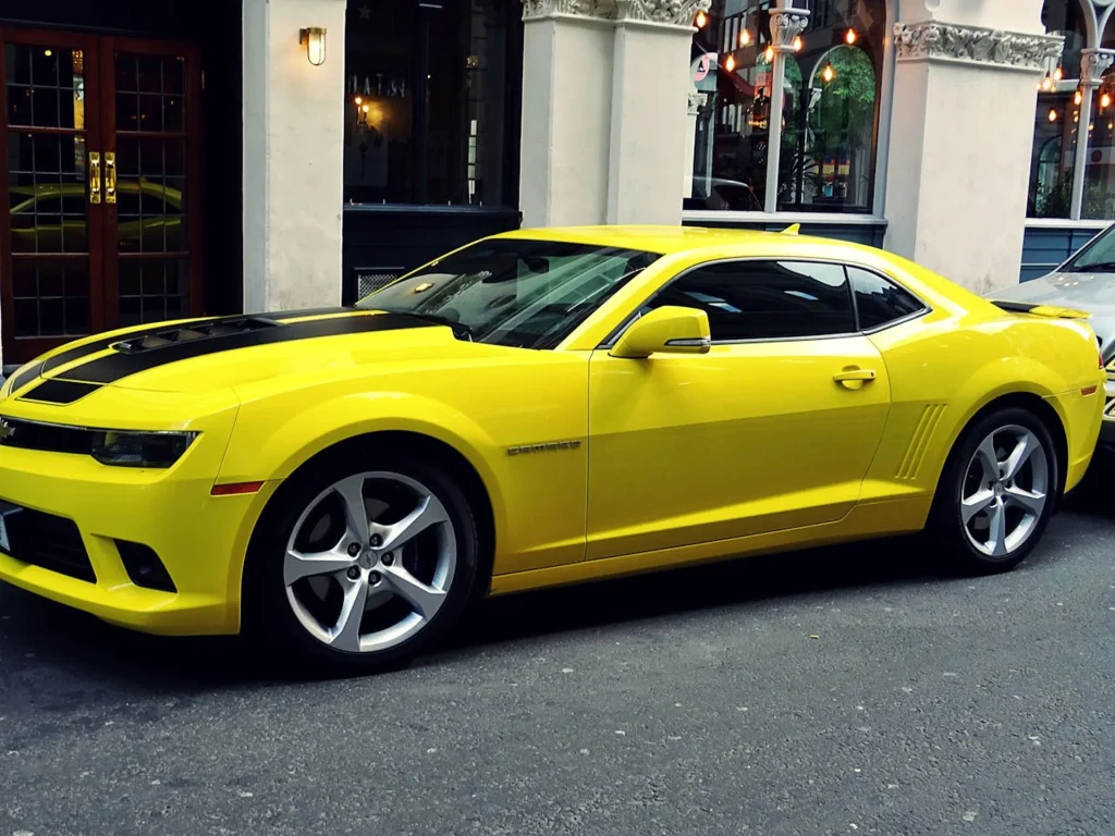 Na zdjęciu znajduje się żółty Chevrolet Camaro zaparkowany na miejskiej ulicy przed eleganckim budynkiem. Samochód ma charakterystyczne, agresywne linie nadwozia oraz czarne pasy wyścigowe na masce, co dodaje mu sportowego wyglądu. W tle widać stylowe okna budynku z metalowymi kratami oraz lampy, co nadaje scenerii eleganckiego i nowoczesnego charakteru. Auto jest w doskonałym stanie, a jego jaskrawy kolor przyciąga uwagę przechodniów.