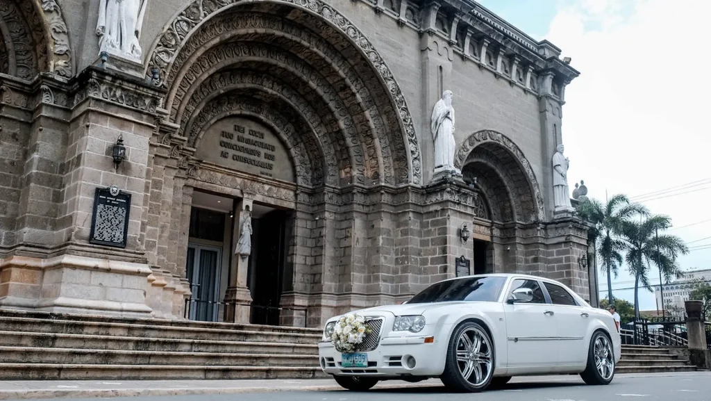 Na zdjęciu znajduje się biały samochód Chrysler 300C, zaparkowany przed imponującą, zabytkową katedrą. Samochód jest ozdobiony bukietem kwiatów na przedniej masce, co sugeruje, że może być używany na okazje ślubne. Chrysler 300C charakteryzuje się eleganckim, nowoczesnym designem, z dużymi, chromowanymi felgami i masywnym grillem. W tle widać bogato zdobione, łukowe wejście do katedry z rzeźbami świętych oraz detale architektoniczne, które podkreślają monumentalny charakter budowli. Roślinność i palmy wokół katedry dodają egzotycznego akcentu do sceny.