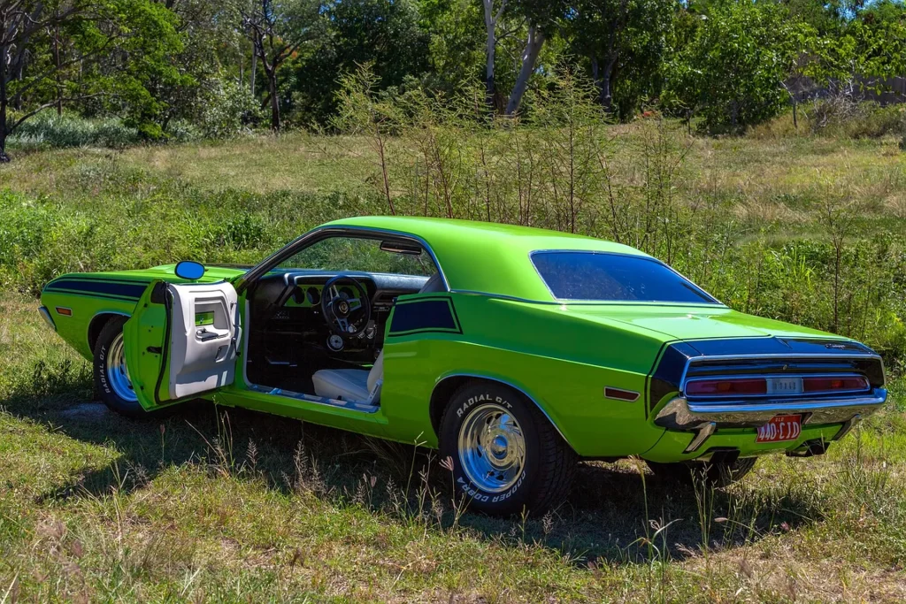 Na zdjęciu znajduje się klasyczny Dodge Challenger w jaskrawozielonym kolorze, zaparkowany na trawie w otoczeniu zieleni. Drzwi kierowcy są otwarte, ukazując wnętrze samochodu. Pojazd ma charakterystyczne dla lat 70-tych linie nadwozia i detale, co podkreśla jego kultowy wygląd. W tle widać drzewa i krzewy, co dodaje zdjęciu naturalnego uroku.