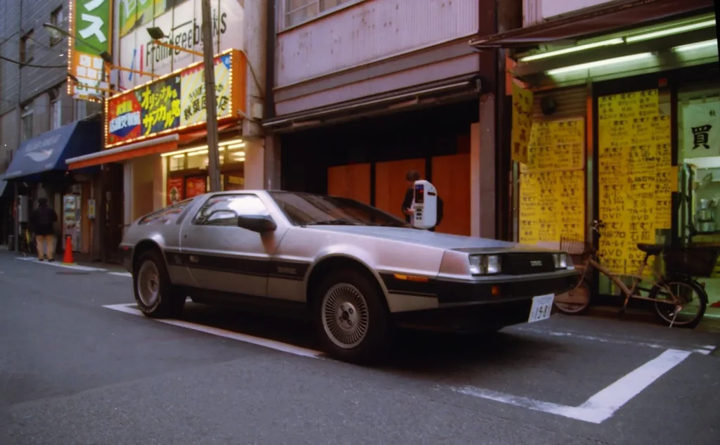 Na zdjęciu widzimy klasyczny DeLorean DMC-12 zaparkowany na ulicy w miejskiej dzielnicy Japonii. Samochód wyróżnia się swoją ikoniczną, stalową karoserią i futurystycznym designem. Ulica jest otoczona budynkami z japońskimi znakami i neonami, co nadaje scenie typowy dla Japonii, tętniący życiem charakter. W tle widać witryny sklepowe oraz osoby korzystające z automatu telefonicznego, co dodaje zdjęciu autentyczności miejskiego życia.