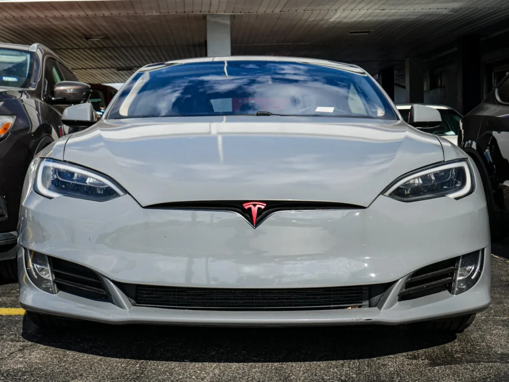 Srebrna Tesla Model S widziana z przodu, zaparkowana na parkingu w otoczeniu innych pojazdów, z charakterystycznym logo na masce i agresywnie stylizowanymi reflektorami.