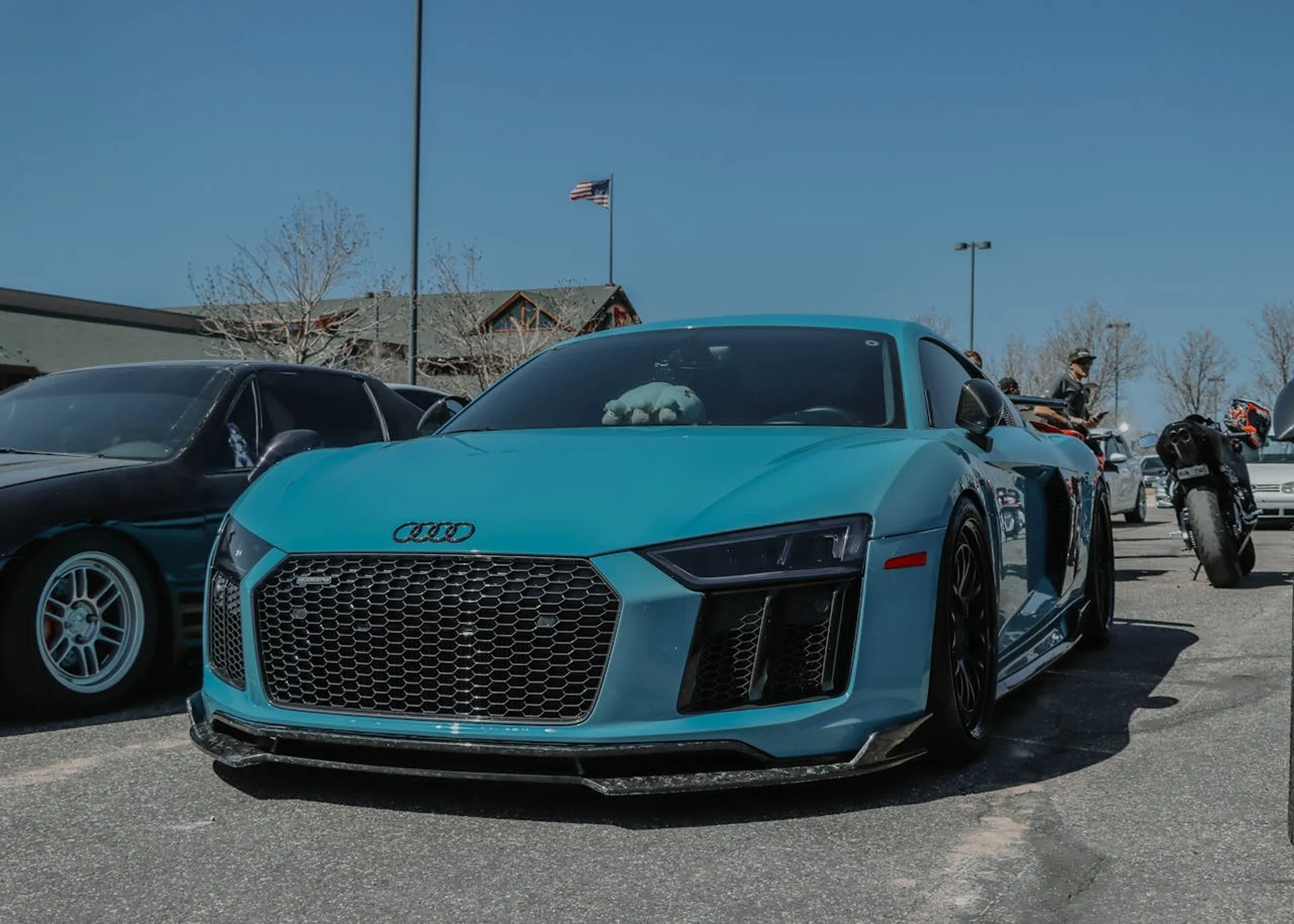 Turkusowe Audi R8 zaparkowane na zlocie samochodowym, obok czarnego auta i motocykla.