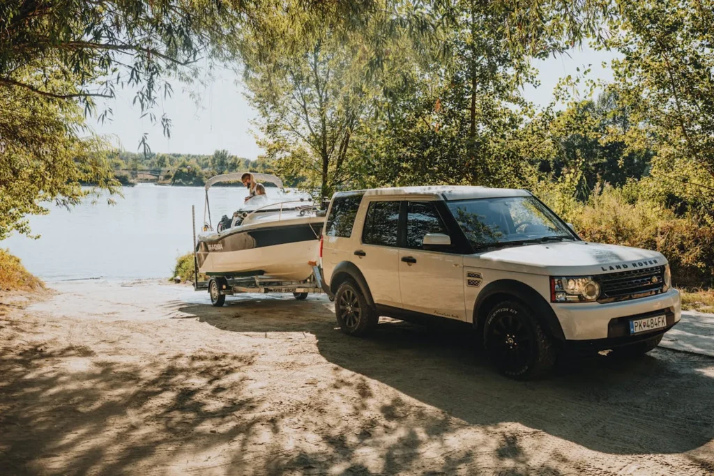 Biały Land Rover Discovery 4 ciągnący motorówkę nad brzegiem rzeki wśród drzew