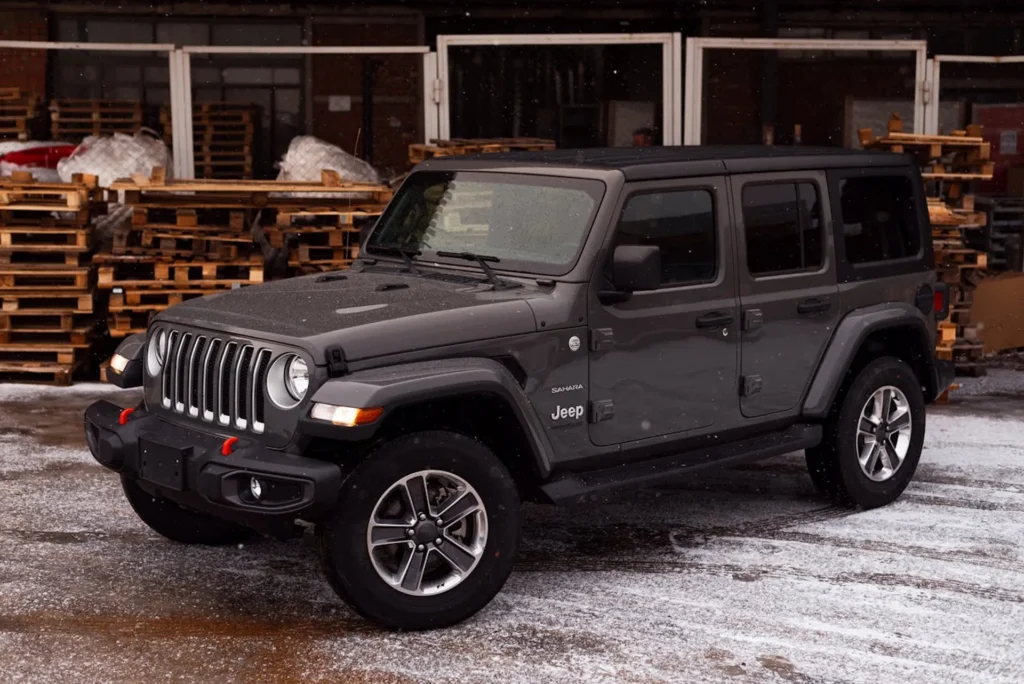 Szary Jeep Wrangler Sahara zaparkowany na placu z paletami, podczas lekkich opadów śniegu