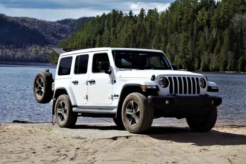 Biały Jeep Wrangler zaparkowany na piaszczystym brzegu jeziora z widokiem na góry i las