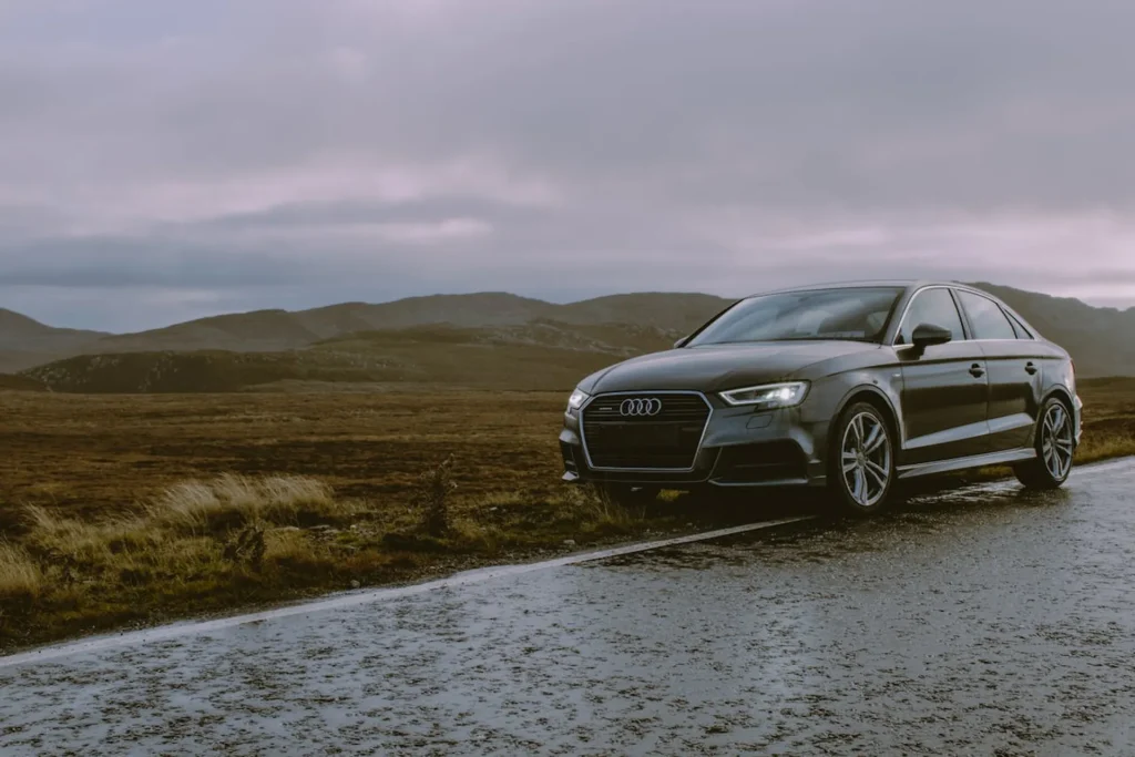 Ciemnoszare Audi na mokrej, górskiej drodze przy pochmurnym niebie.
