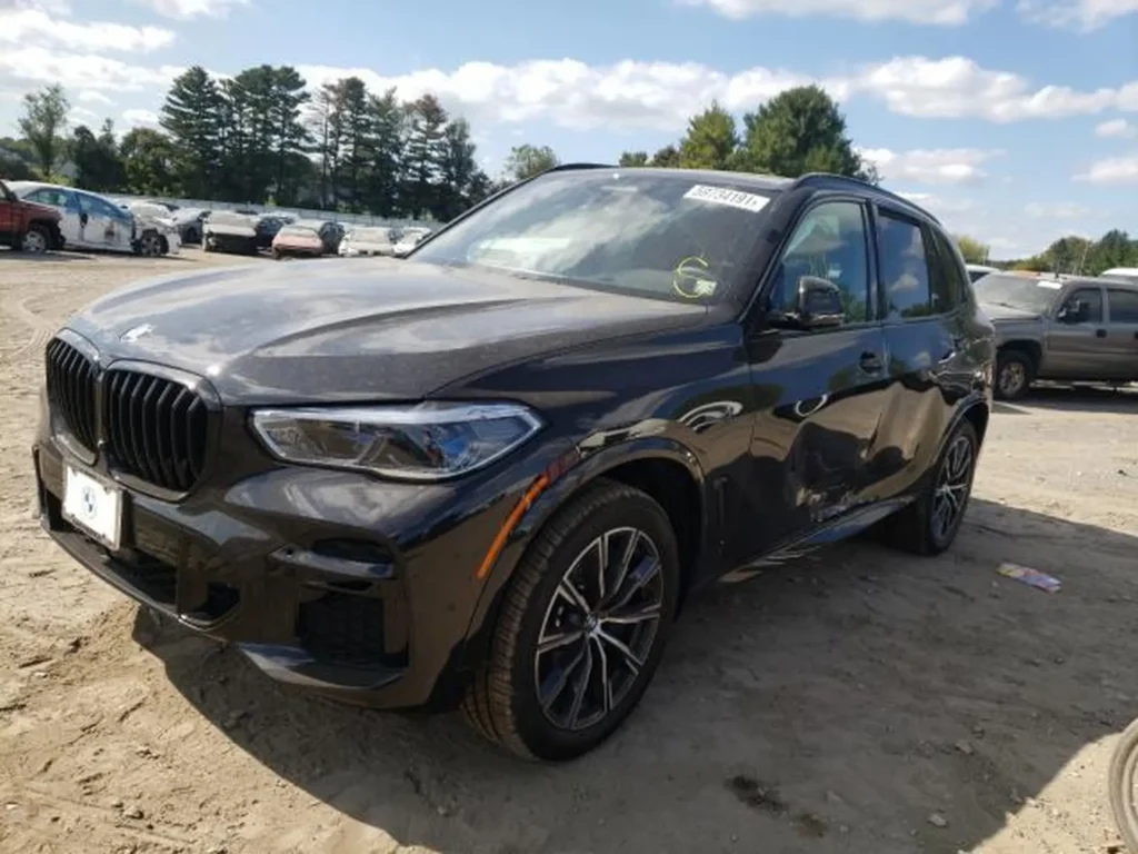 BMW X5 z przodu z prawej, widoczne wgniecenie na prawych drzwiach, parking żwirowy.