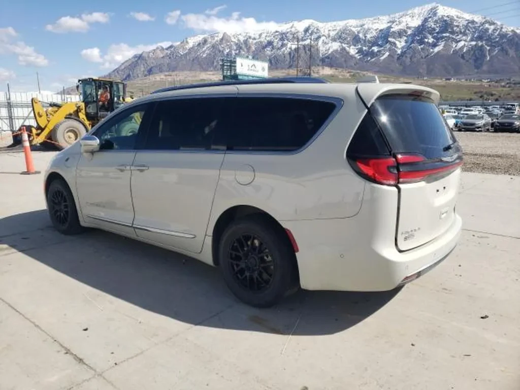 Jasny Chrysler Pacifica z czarnymi felgami, widok z tyłu i boku, zaparkowany na placu z ośnieżonymi górami w tle.