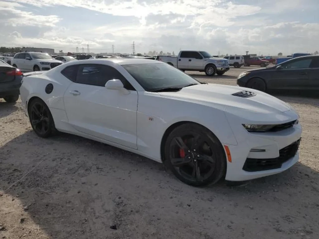Biały Chevrolet Camaro zaparkowany na żwirowym placu wśród innych aut