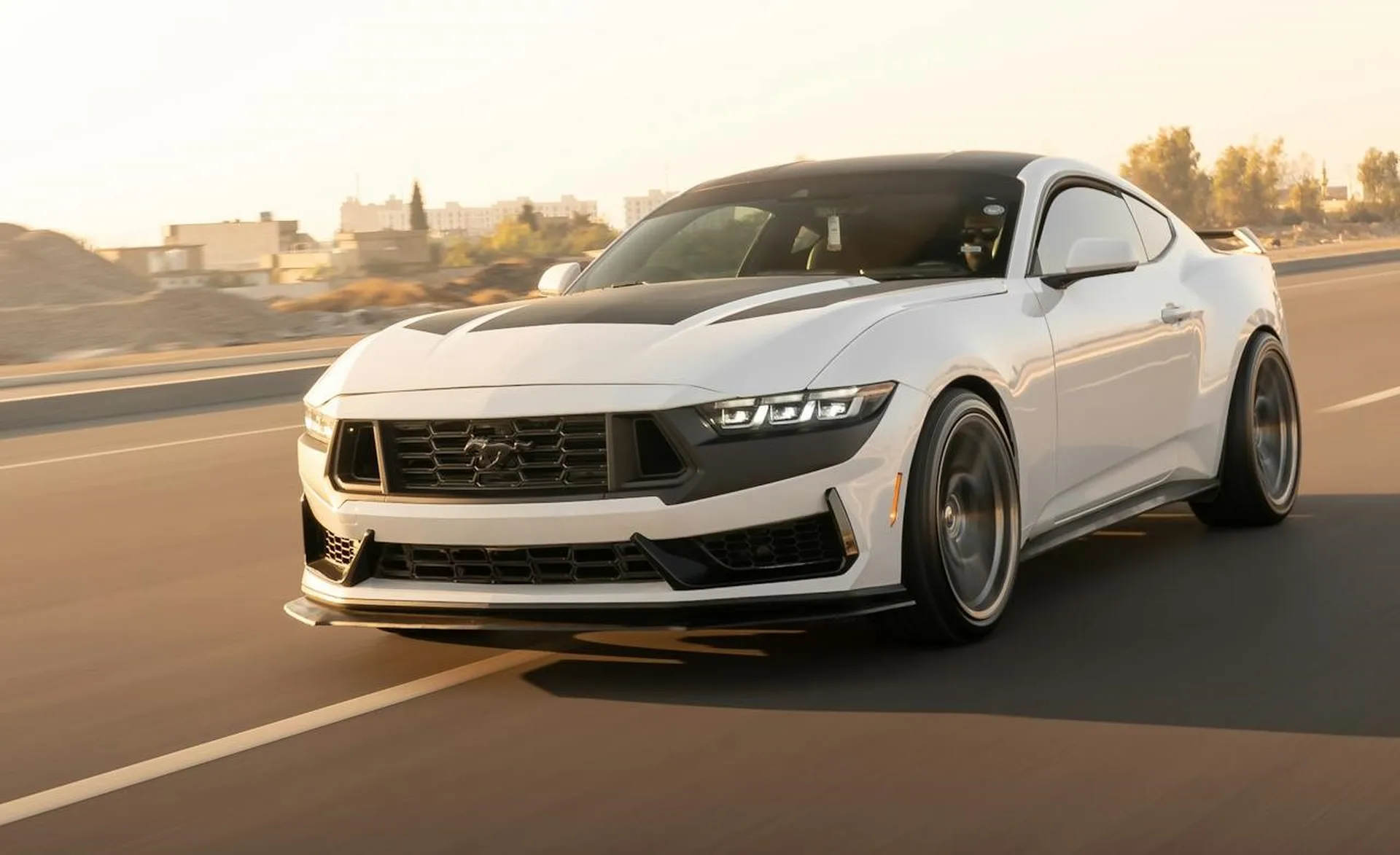 Biały Ford Mustang Dark Horse w ruchu na autostradzie o zachodzie słońca, z agresywnym przodem i sportową sylwetką.